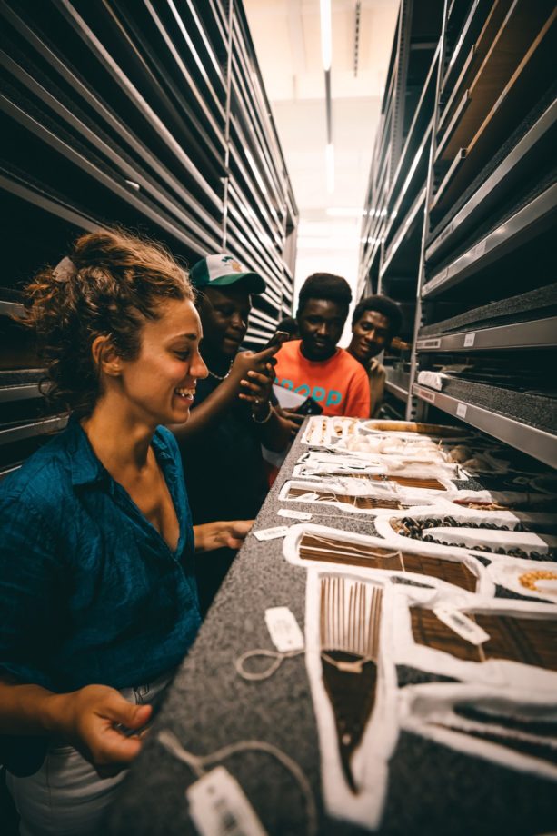 Four individuals stand in an isle between tall shelves in a museum depot and examine labeled items on a shelf.